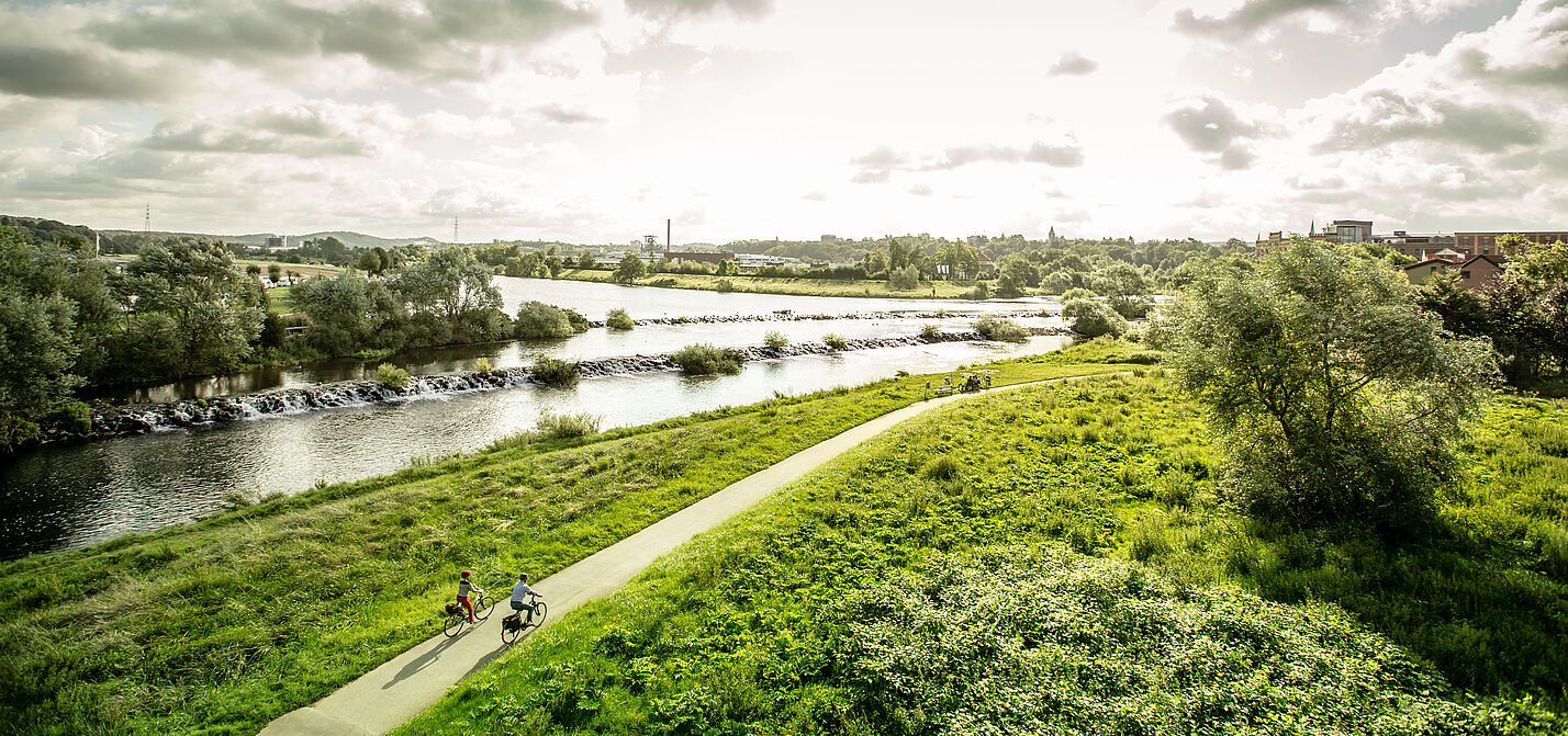 Panoramablick aus der Vogelperspektive auf den Ruhrtalradweg entlang der Ruhr, zwei Radfahrende fahren der Nachmittagssonne entgegen.